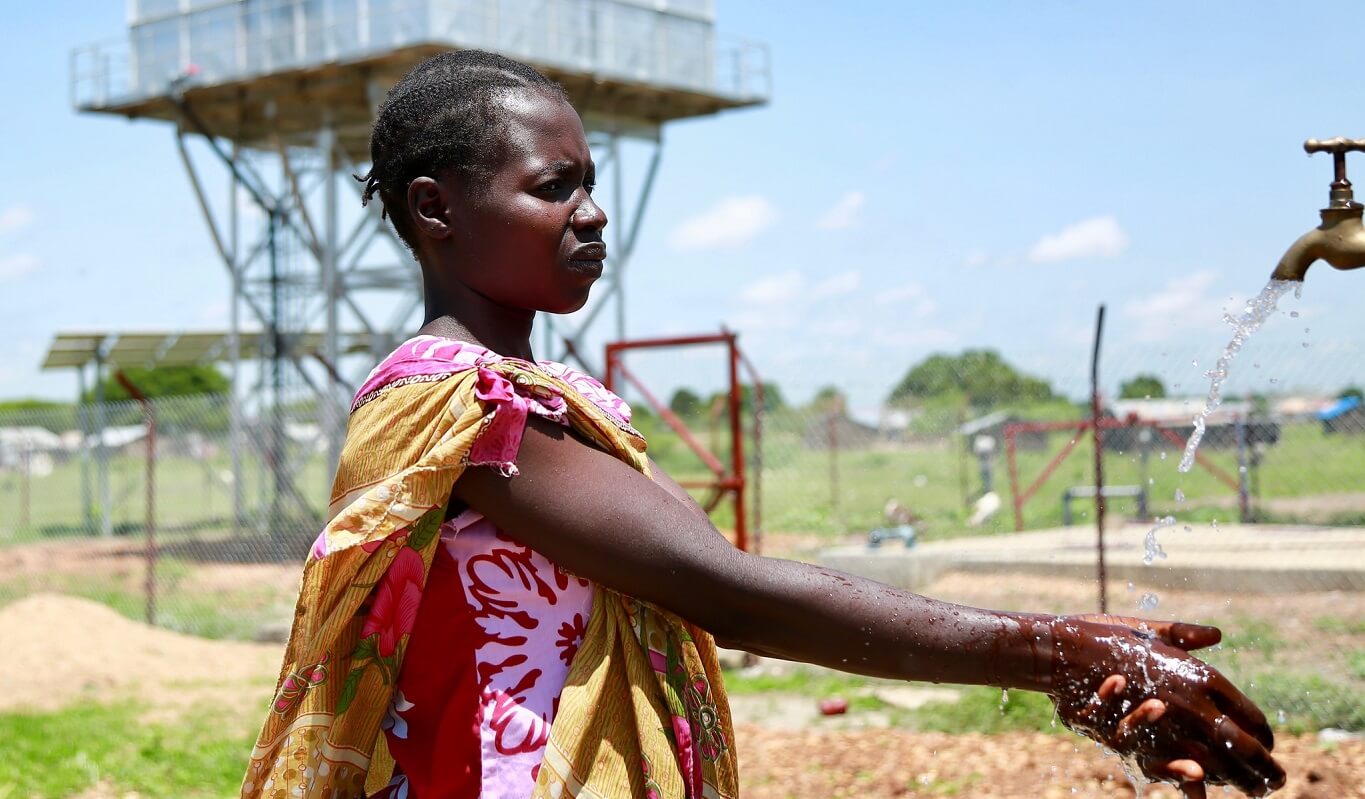 A black woman in brightly coloured clothing holds out her hands under a water tap to wash them. The water tower can be seen in the background.