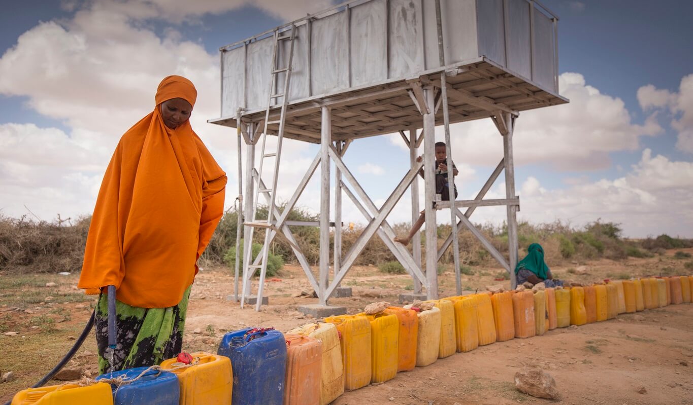 A woman in an orange head covering and green dress stands over a long line of water cans, filling them with a hose.