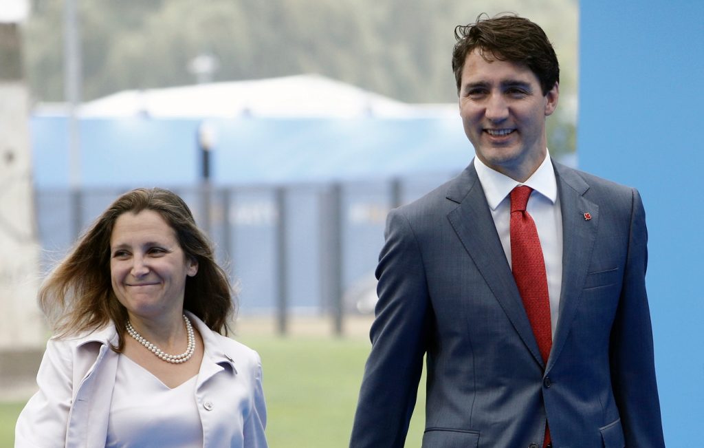 On the left of the image, The Honourable Chrystia Freeland walks and smiles while wearing a white jacket and dress, and on the right of the image PM Justin Trudeau walks and smiles wearing a grey suit, white button-down shirt and a red tie.