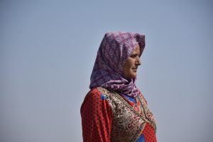 Woman in red patterned dress and purple hijab in blue sky background looking towards the right.