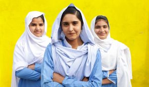Three girls in white headscarves and blue dresses against a bright yellow wall cross their arms and smile at the camera