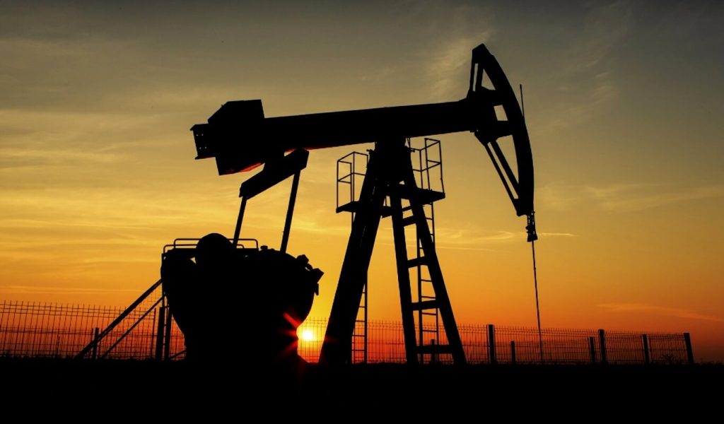 In the foreground, we see industrial oil drilling equipment in an open field and in the background there is a colourful sunset in shades of orange, yellow and red.