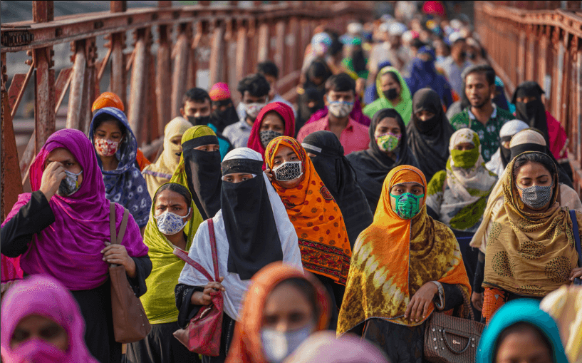A crowd consisting of people of various genders and skin tones are walking towards us across a bridge during the day, in an urban environment, wearing a variety of colourful clothes, face masks, and religious garb.