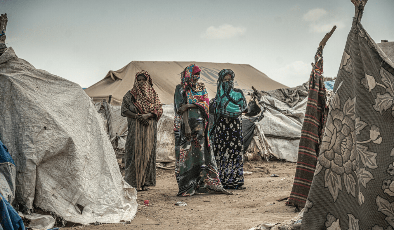 Three women wearing colourful hijabs stand outside, surrounded by white, makeshift tents against a cloudy sky. Beneath them the ground is brown.