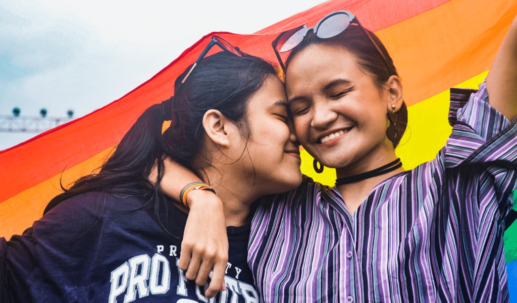 Two young Brown women embrace each other. The woman on our left is pressing her nose agains the cheek of the woman in our right whose eyes are closed while she smiles and raises one of her arms. Behind them is a rainbow flag.