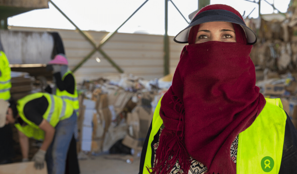 Two people wearing Oxfam branded fluorescent green vests and work gloves separate card board to our left. To our right a woman wearing a dark red headscarf covering her face looks at the distance. She's also wearing the Oxfam branded vest.