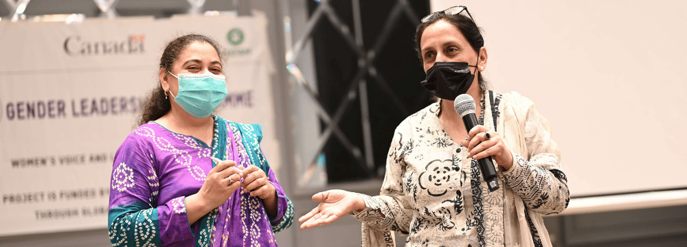 Two women wearing medical facemasks are standing up and holding microphones.