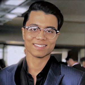 A young person with black rimmed glasses and a navy blue suit, smiling.