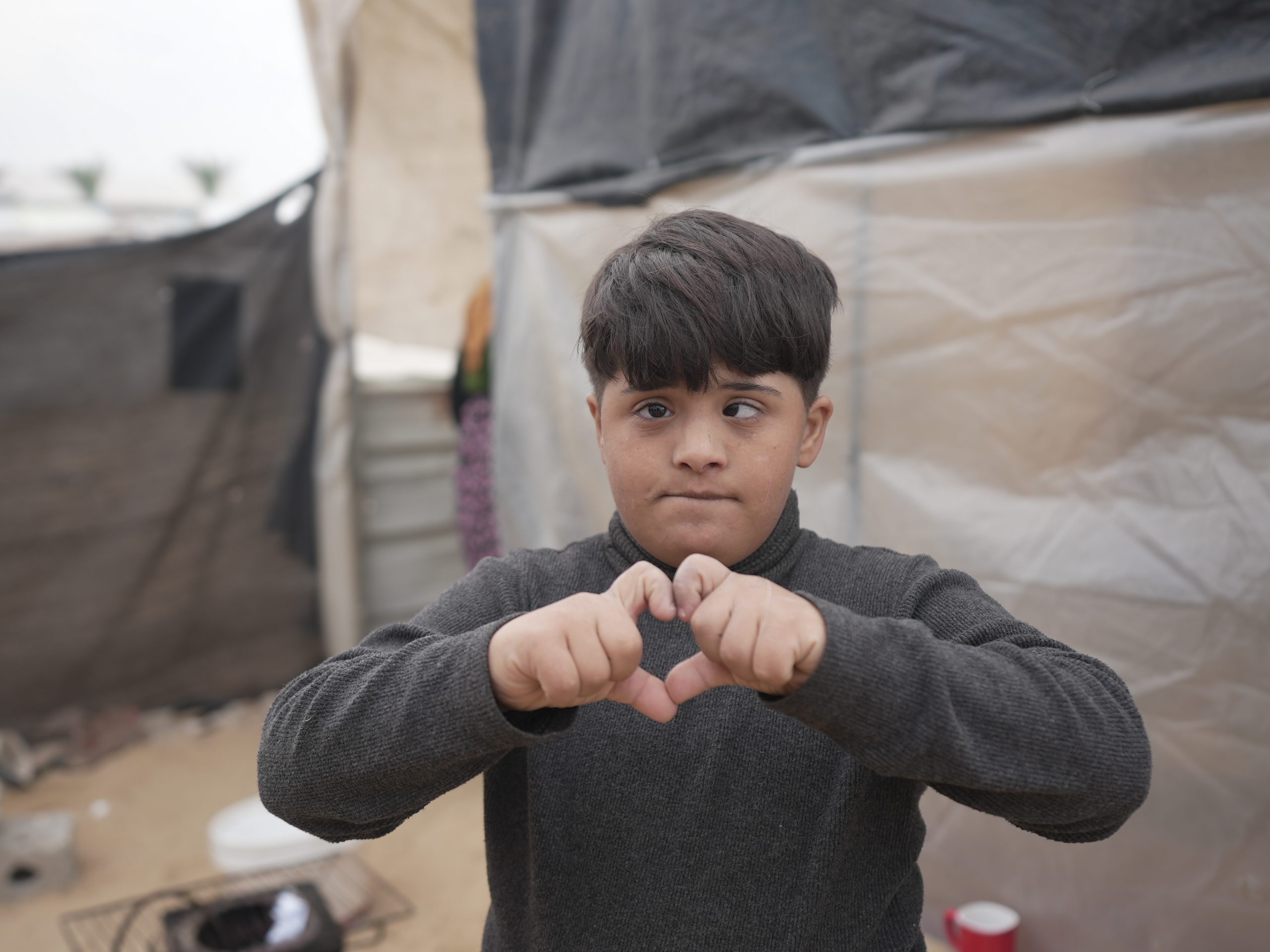 Mutaz's son with Down syndrome makes a love sign while standing in front of his family tent in Al Mawasi.