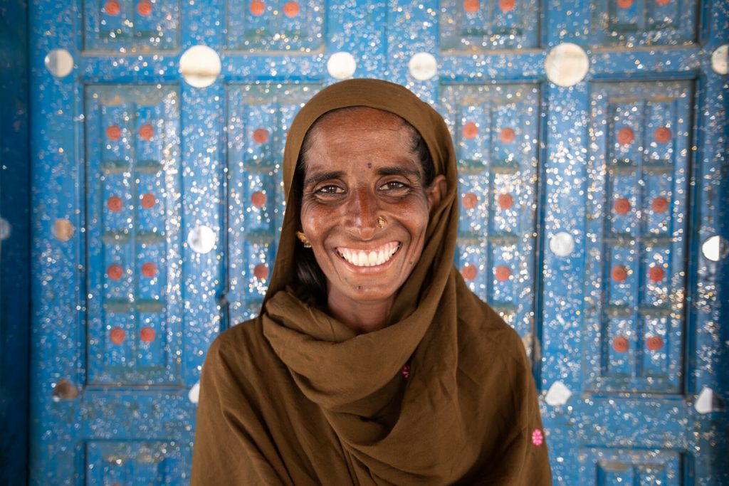 Woman wearing a brown headscarf smiles directly at camera while standing in front of a blue door.