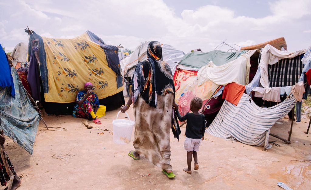 A woman and her young girl walk through a camp of makeshift tents, the ground is wet and muddy, and the child is walking barefoot.