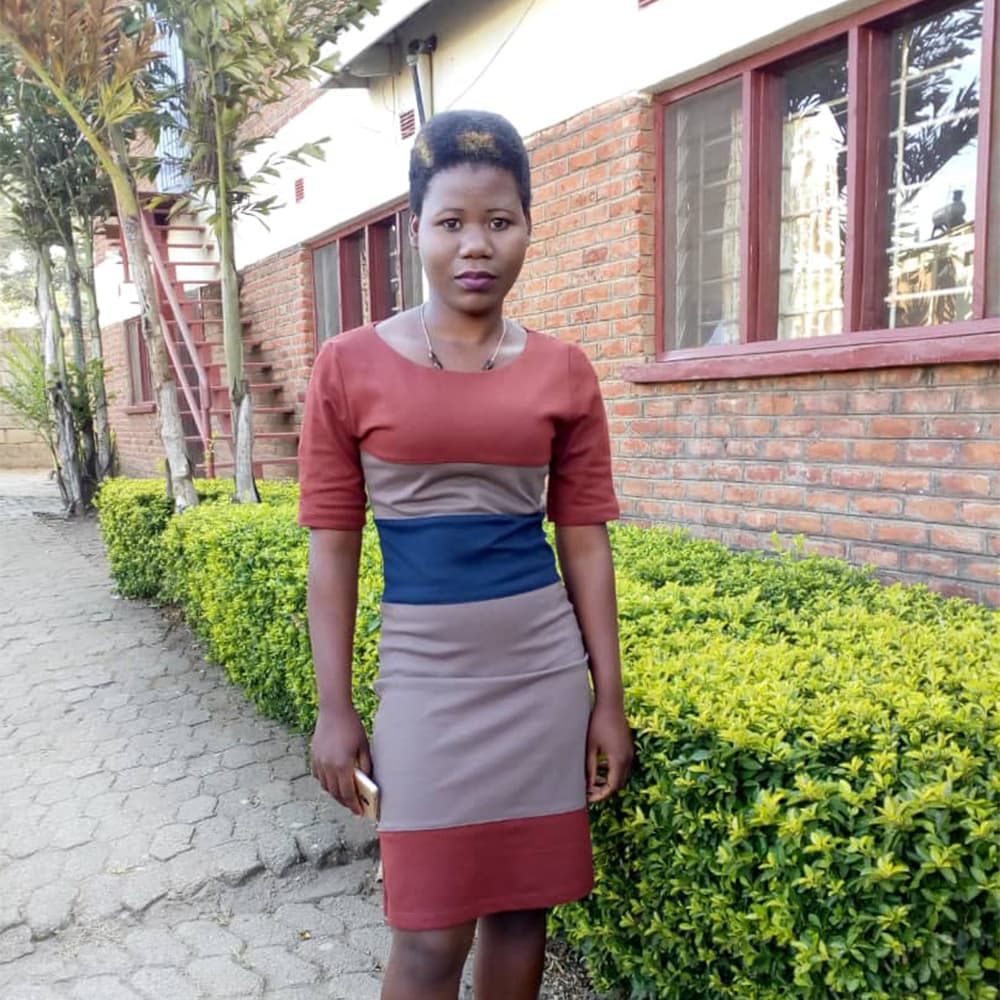 A young woman in Malawi, with a short afro hairstyle and wearing a striped red and blue dress, looking at the camera while standing outside of a red brick building surrounded by bright green shrubs.