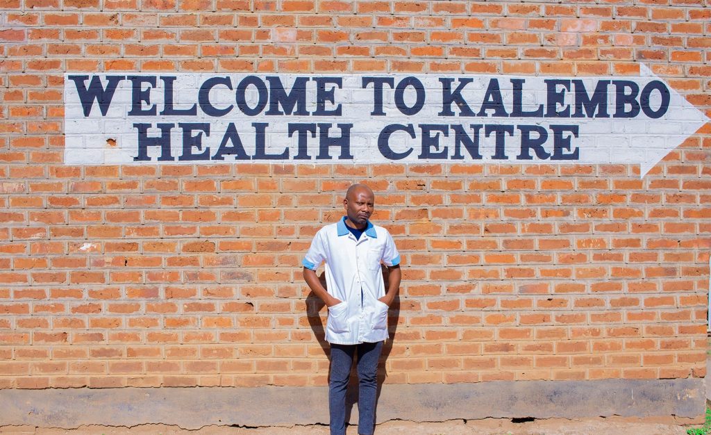 A young Malawian man, wearing a blue and white medical shirt, standing in front of a brick wall adorned with a painted sign that read, "Welcome to Kalembo Heath Centre."