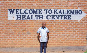 A young Malawian man, wearing a blue and white medical shirt, standing in front of a brick wall adorned with a painted sign that read, "Welcome to Kalembo Heath Centre."