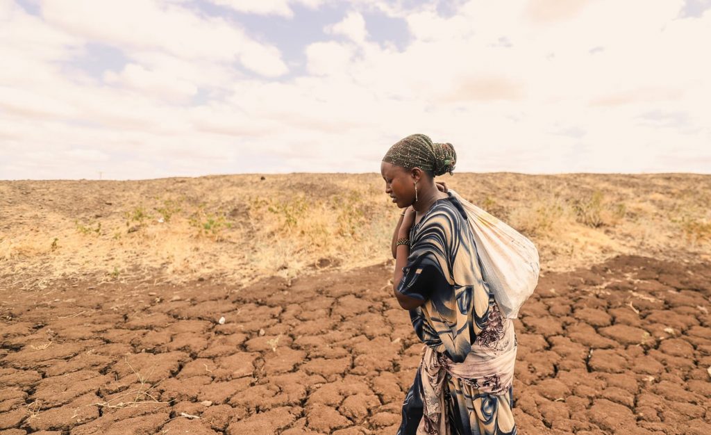 A woman in a headscarf and a white back over her shoulder walks through an arid, drought-stricken place with cracked earth.