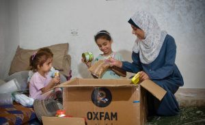 A Palestinian woman in a headscarf, is opening a box of canned food and cereals with her two young daughters in a bare room.