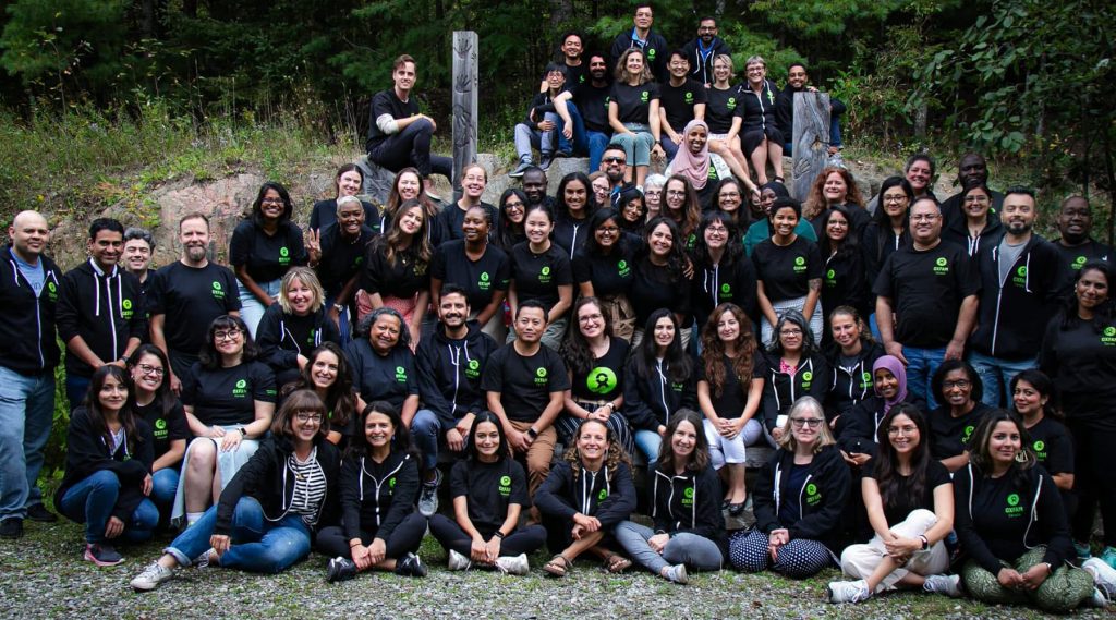 A team of dozens of individuals from diverse backgrounds sitting for a group photo in an outdoor, forested setting.