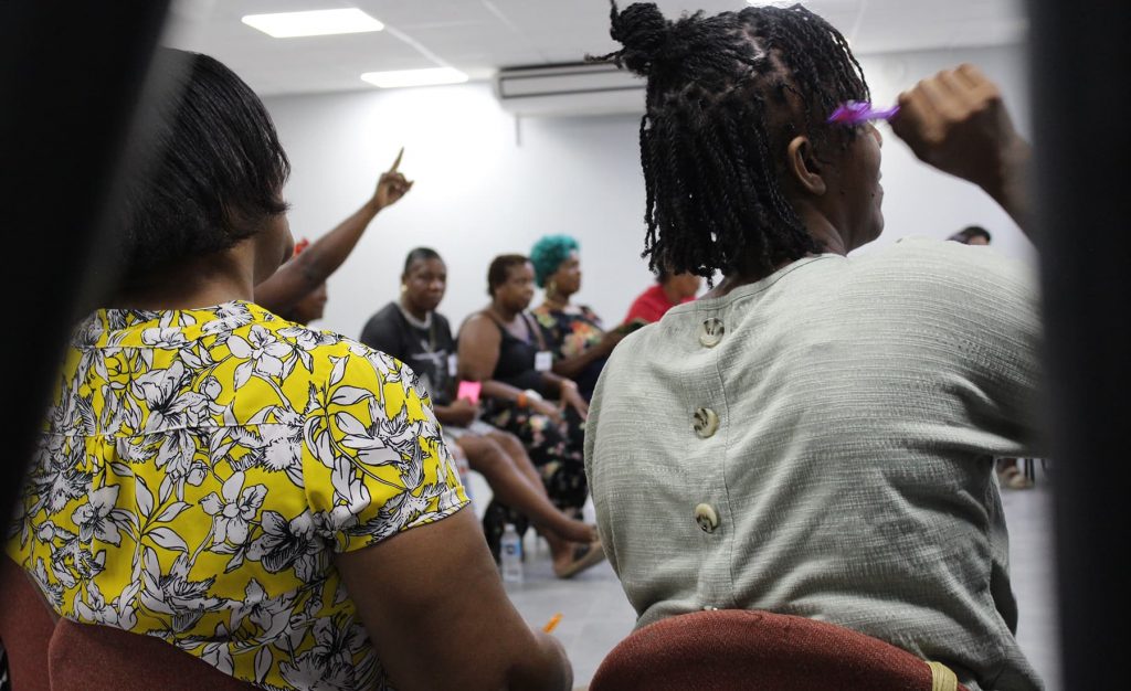 A group of Jamaican women sitting in a circle during a workshop in a grey classroom, the women are raising their hands to answer questions.