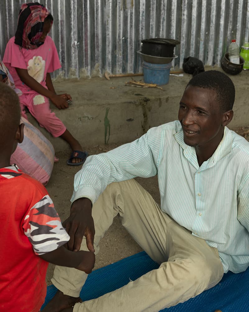 A young man sitting on a blue mat on the ground, talking to a young boy in a red shirt.