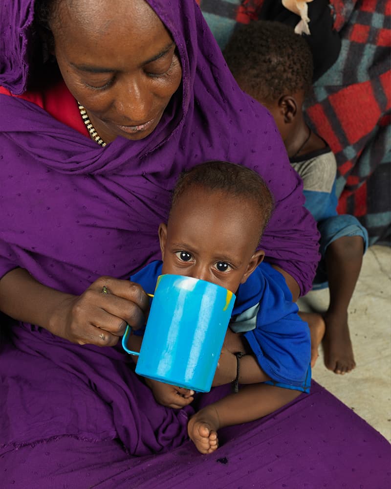 A woman in a purple headscarf feeding a small boy from a blue mug.