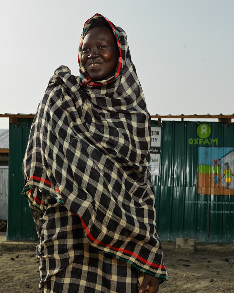 A woman wearing a checkered headscarf and smiling at the camera in front of a green, tin shelter with the Oxfam logo on it.