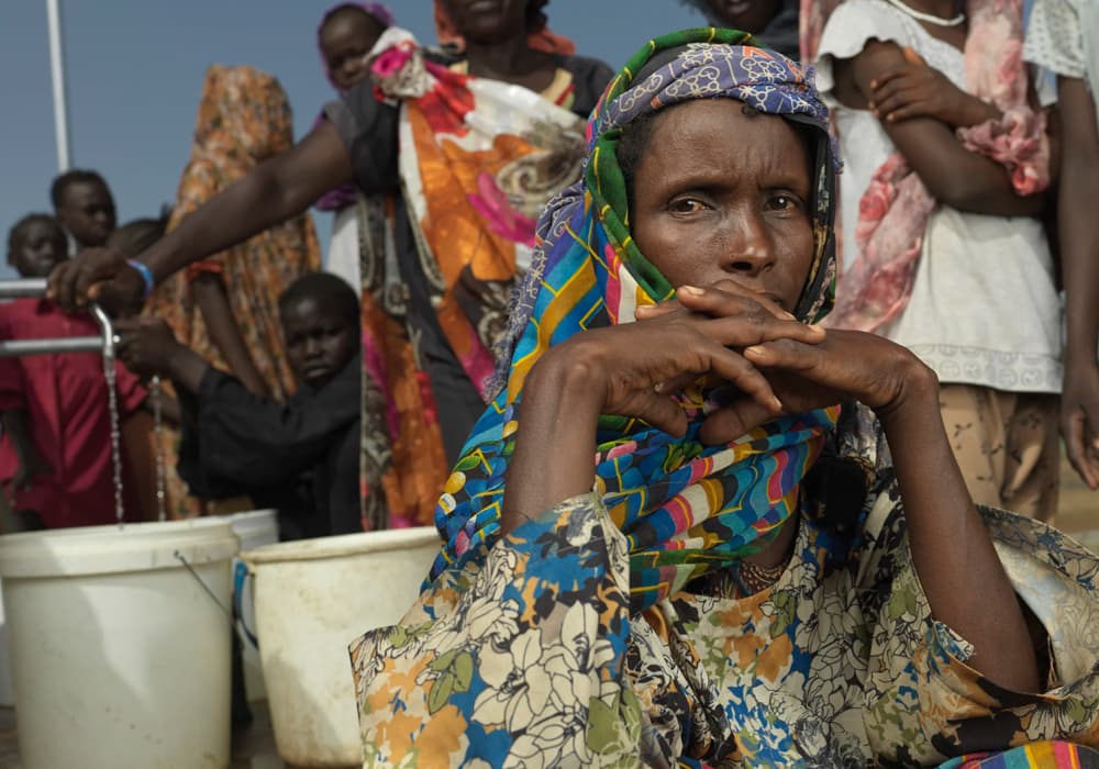 A woman sitting in front of a makeshift water tap and a line of people with an inquisitive look.