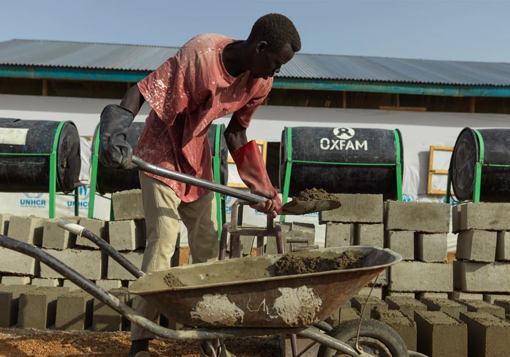 A young man building a wall from bricks and concrete in front of a large shelter with Oxfam logos on it.