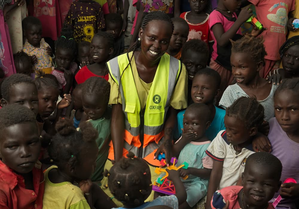 A young woman wearing an Oxfam vest, surrounded by young girls and boys, all smiling at the camera.