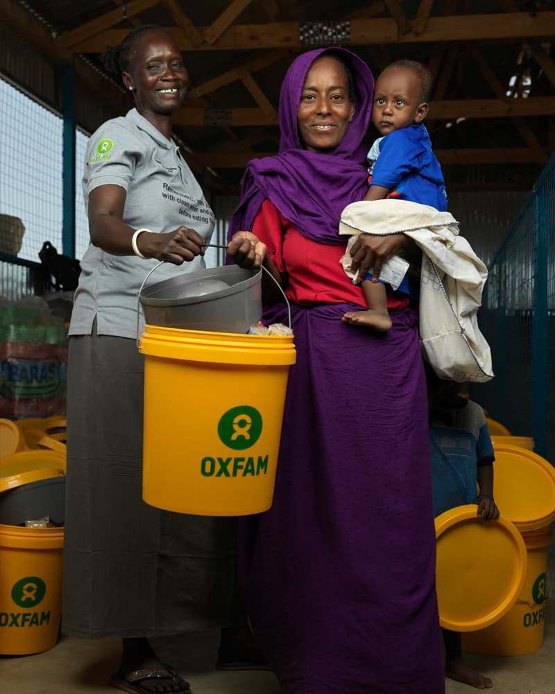 A women in a purple headscarf and a woman in an Oxfam uniform holding a yellow and green bucket.