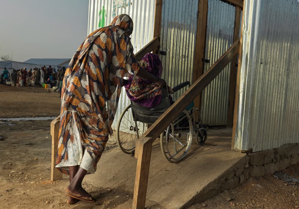 A woman wheeling an older woman in a wheelchair up a ramp into a tin shelter.