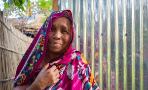 A woman wearing a vibrant, pink floral headscarf smiling and standing in front of a shiny tin wall, with a fence and trees in the background.