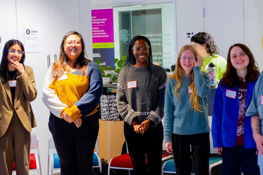 Five young people of varying ethnicities and identities standing in an office-looking room and smiling at the camera.