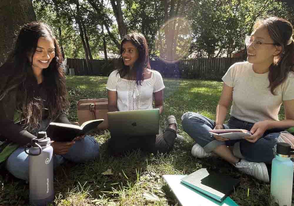 Three university-aged women are holding a meeting outdoors.